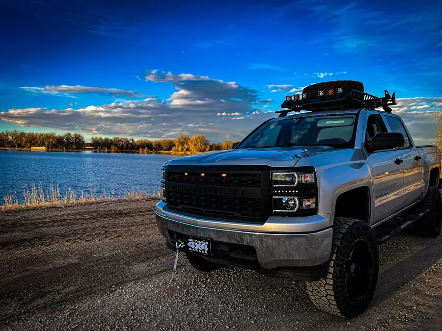 Lifted silver Chevy Silverado with a roof rack, light pods and a blacked-out grille beside a lake